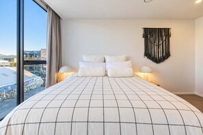 A hotel room with a large bed, two bedside lamps, a wall-mounted macrame piece, and a view of buildings through a large window.