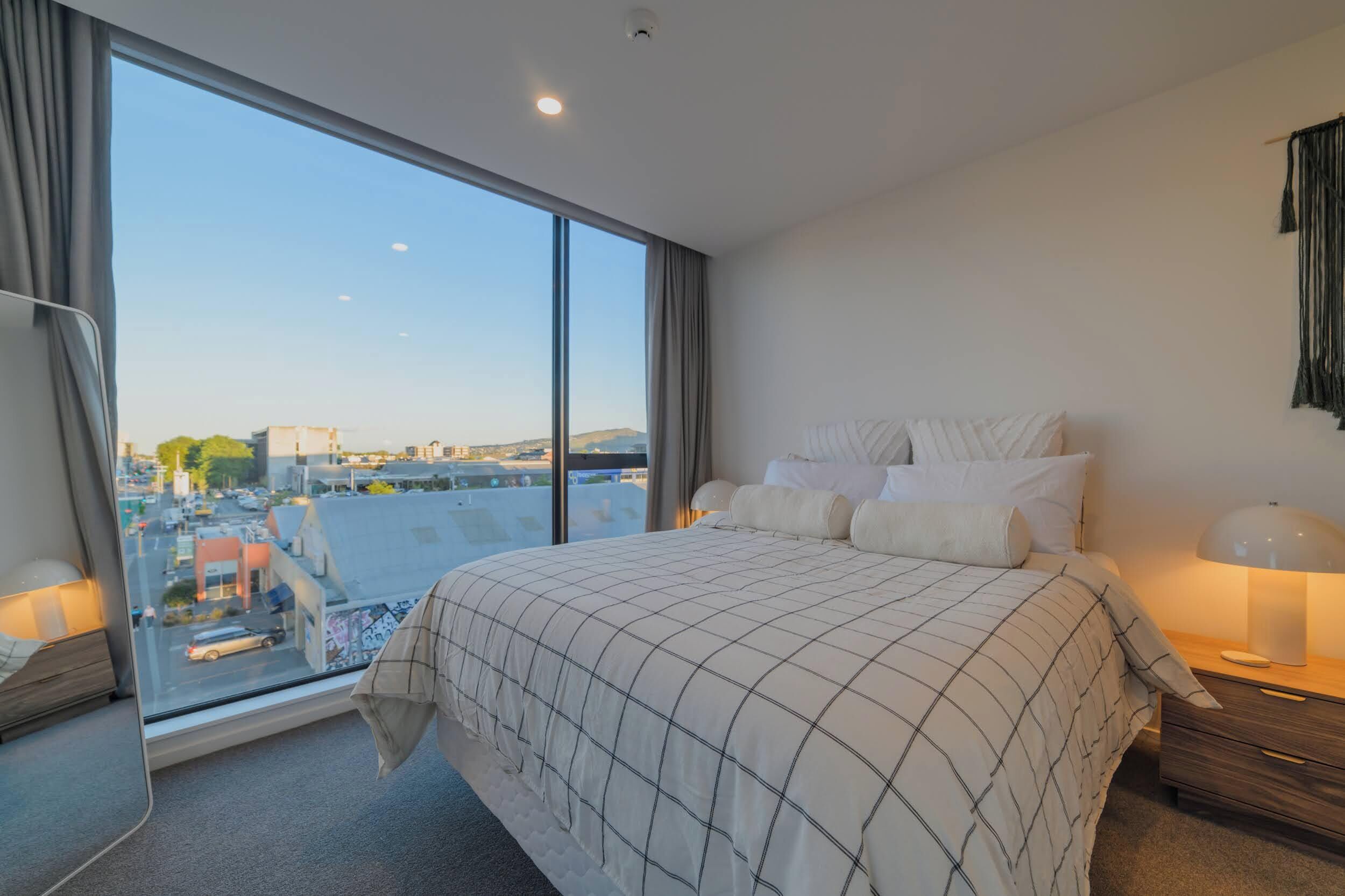 A modern hotel room with a large window, a neatly made bed, a bedside lamp, and a view of the cityscape.