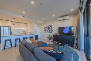 Una sala de estar moderna con un sofá, un comedor con taburetes, un televisor y una cocina al fondo.