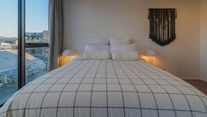 A bedroom with a large bed, two bedside lamps, a wall-mounted tapestry, and a view of buildings through a large window. - Cityside Comfort!  Modern 2BR, 2BA with Parking (Christchurch)
