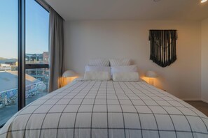Ein Schlafzimmer mit einem großen Bett, zwei Nachttischlampen, einem Wandteppich und Blick auf Gebäude durch ein großes Fenster.