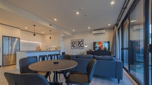 Een moderne woonkamer met een grijze bank, een eettafel met stoelen en een keuken met een koelkast en een spoelbak.