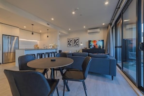 Una sala de estar moderna con un sofá gris, una mesa de comedor con sillas y una cocina con refrigerador y lavaplatos.