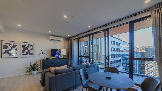 A modern living room with a sofa set, a dining table, and chairs. There are large windows with a view of buildings outside. - Cityside Comfort!  Modern 2BR, 2BA with Parking (Christchurch)