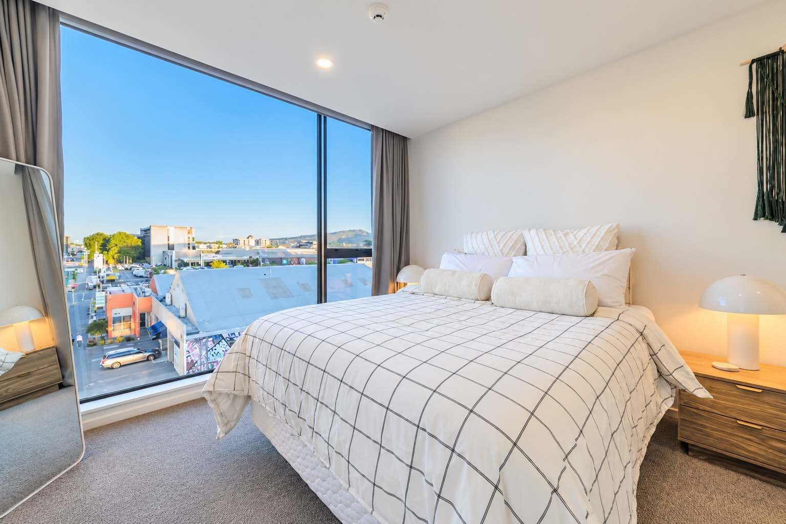 Un dormitorio con una cama grande, una mesita de noche, una lámpara y vistas de la ciudad a través de amplios ventanales.