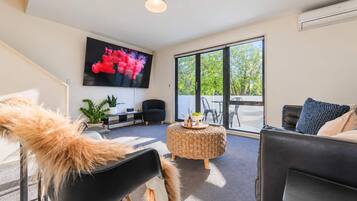 A modern living room with a large TV, comfortable seating, a small round table, and a view through large windows.