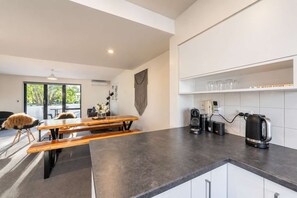A modern kitchen with a black countertop, a wooden dining table with benches, and a coffee station with a kettle and coffee maker.