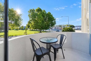 En balkon med et glasbord og to stole, der kigger ud over en grøn græsplæne og en gade.