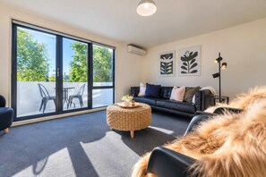 A modern living room with a sofa, a round wicker coffee table, and a large sliding glass door leading to an outdoor seating area.
