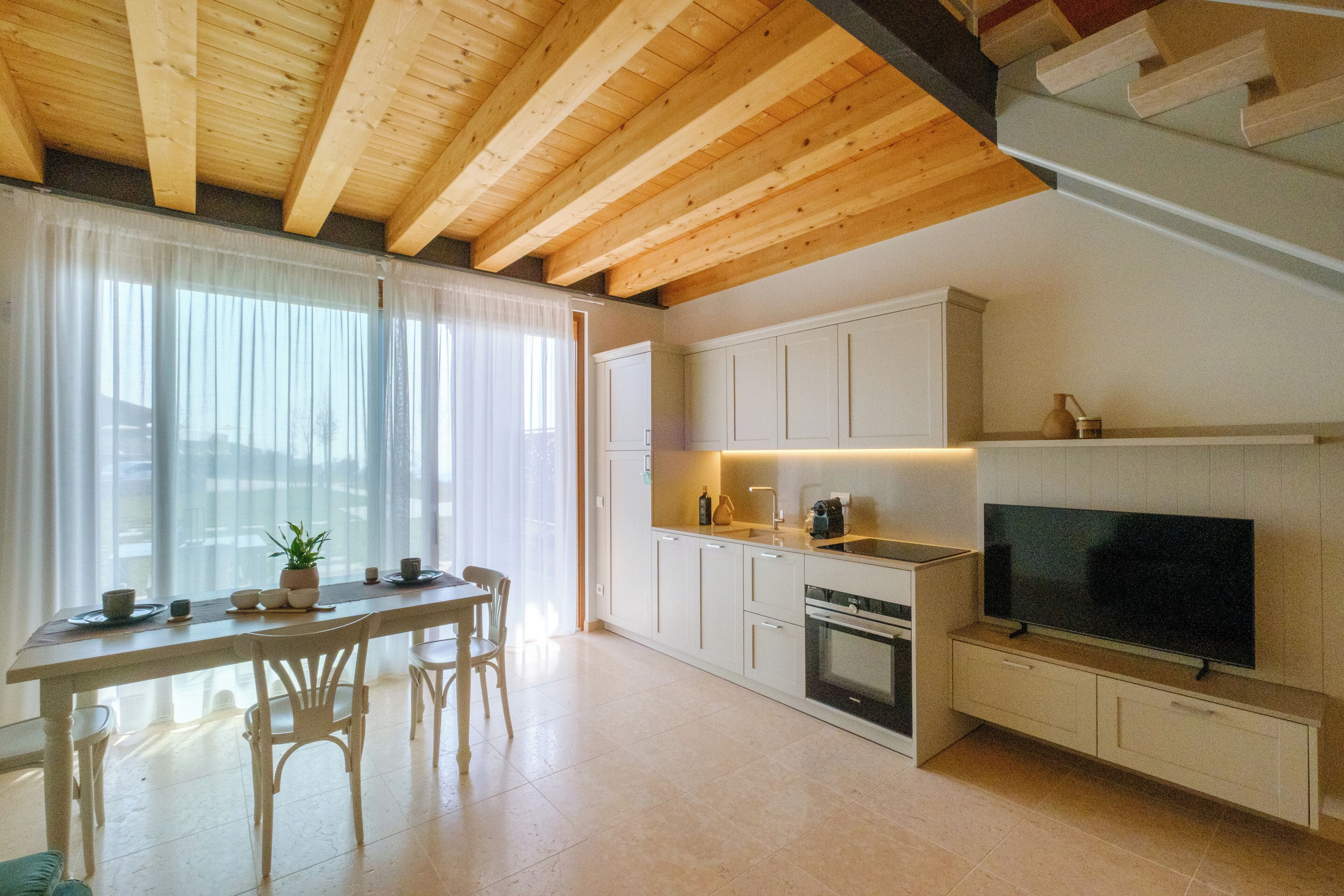 Eine moderne Küche mit Holzbalken, weißen Schränken, einem Essbereich mit Stühlen und einem großen Flachbildfernseher an der Wand.