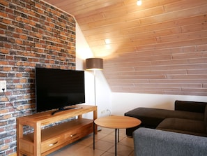 Un salon moderne avec un mur en briques, un plafond en bois, un téléviseur à écran plat posé sur un meuble en bois, une petite table ronde et un canapé gris.