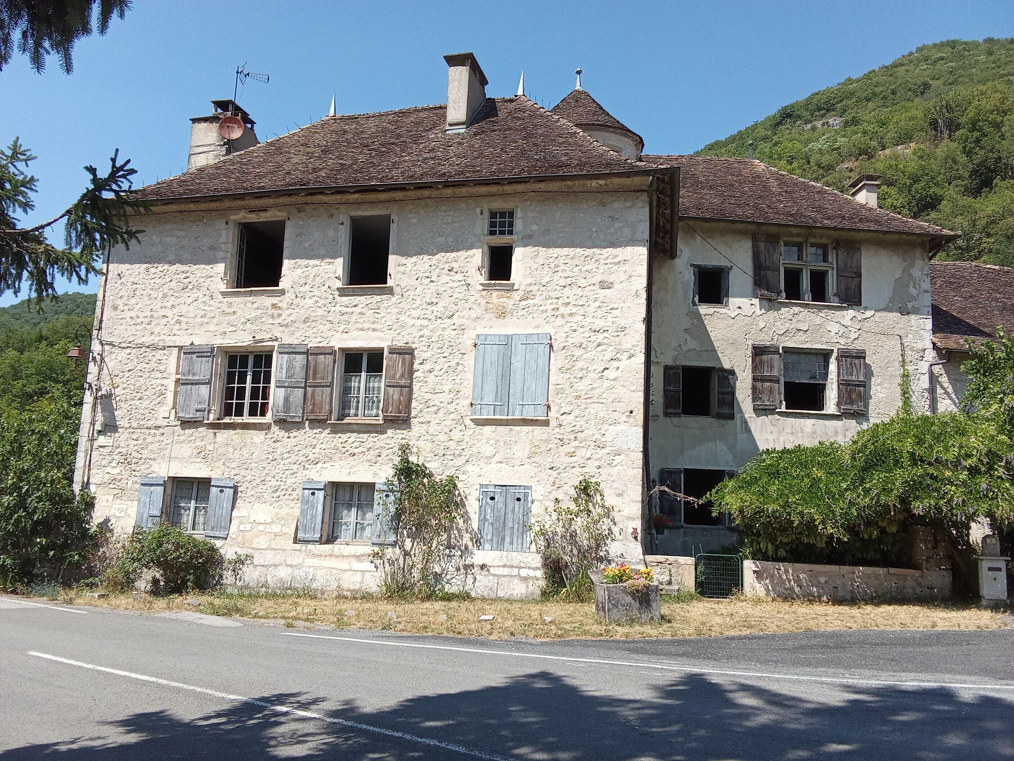 Un bâtiment en pierre à deux étages, doté de nombreuses fenêtres et de volets, entouré de verdure et situé au pied d’une colline.