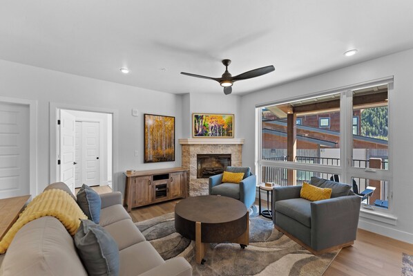 A living room with a fireplace, a ceiling fan, and large windows. - Ski & Stay in Style – Condo Near Lifts w/ Pool, Hot Tubs & Fireplace (Keystone)