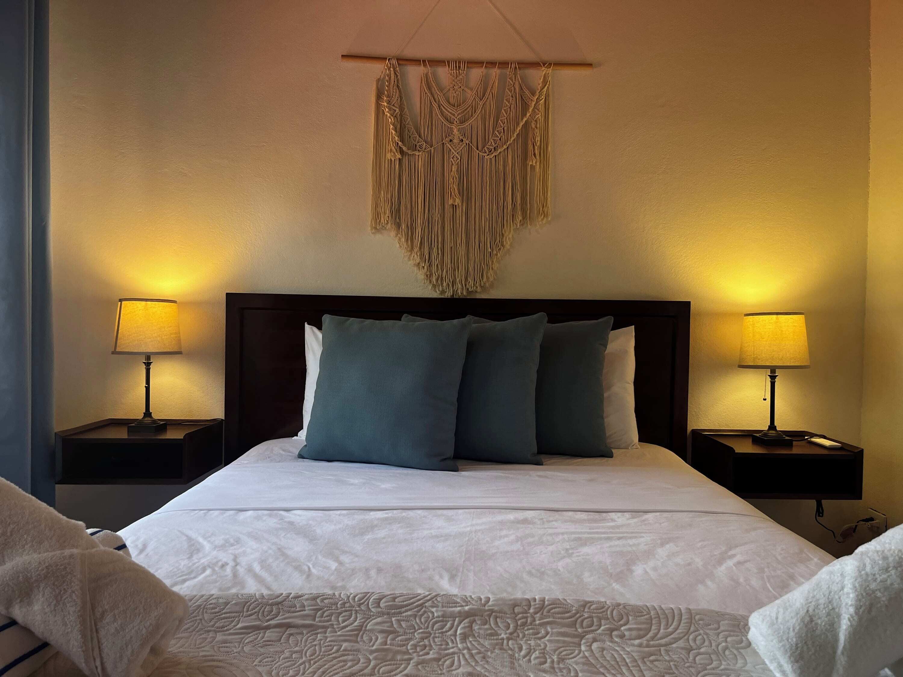 A neatly made bed with white linens and a decorative headboard. There are two bedside tables with lamps, and a macramé wall hanging above the bed.
