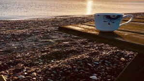 En strandscen med en kopp på en träyta, småstenar och solen nära horisonten.