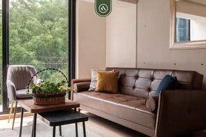 A modern living room with a leather sofa, a chair, a small table with a plant, and a large window with a view of trees. - Brand New 2 BR Condo in Reforma - Queen Beds (Mexico City)