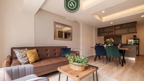 Ein modernes Wohnzimmer mit einer braunen Couch, einem grauen Sessel und einem Couchtisch mit einem Blumenkorb. Im Hintergrund ist eine Küche mit Holzschränken und ein Essbereich mit Stühlen erkennbar.