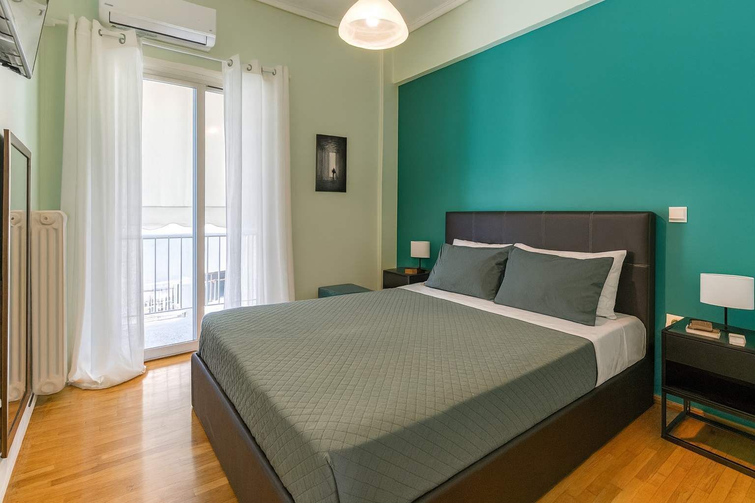 A bedroom with a bed, a nightstand, a window with curtains, and a view of a balcony.