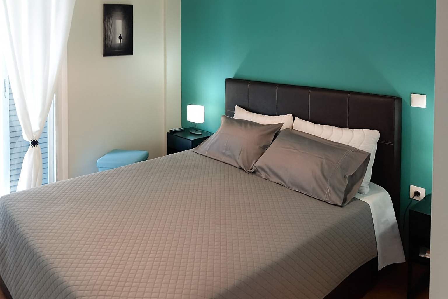 Ein ordentlich bezogenes Bett mit einer gesteppten Tagesdecke, zwei Kissen und einem Kopfteil vor einer türkisblauen Wand. Ein Nachttisch mit Lampe und Steckdose ist ebenfalls zu sehen.