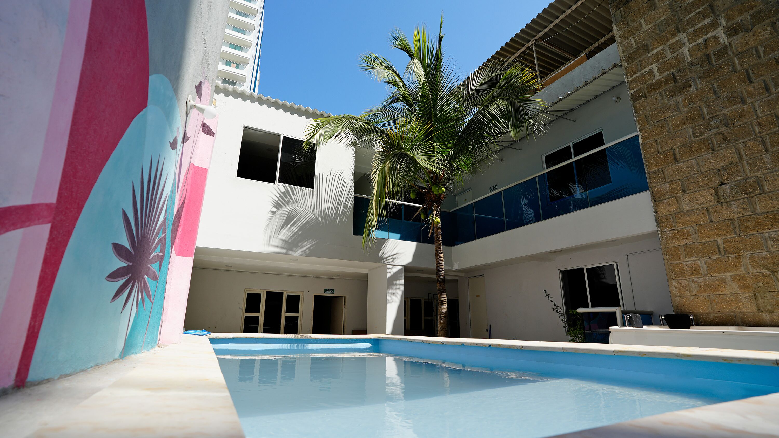 A swimming pool with a palm tree, a building with a balcony, and a mural on the wall.