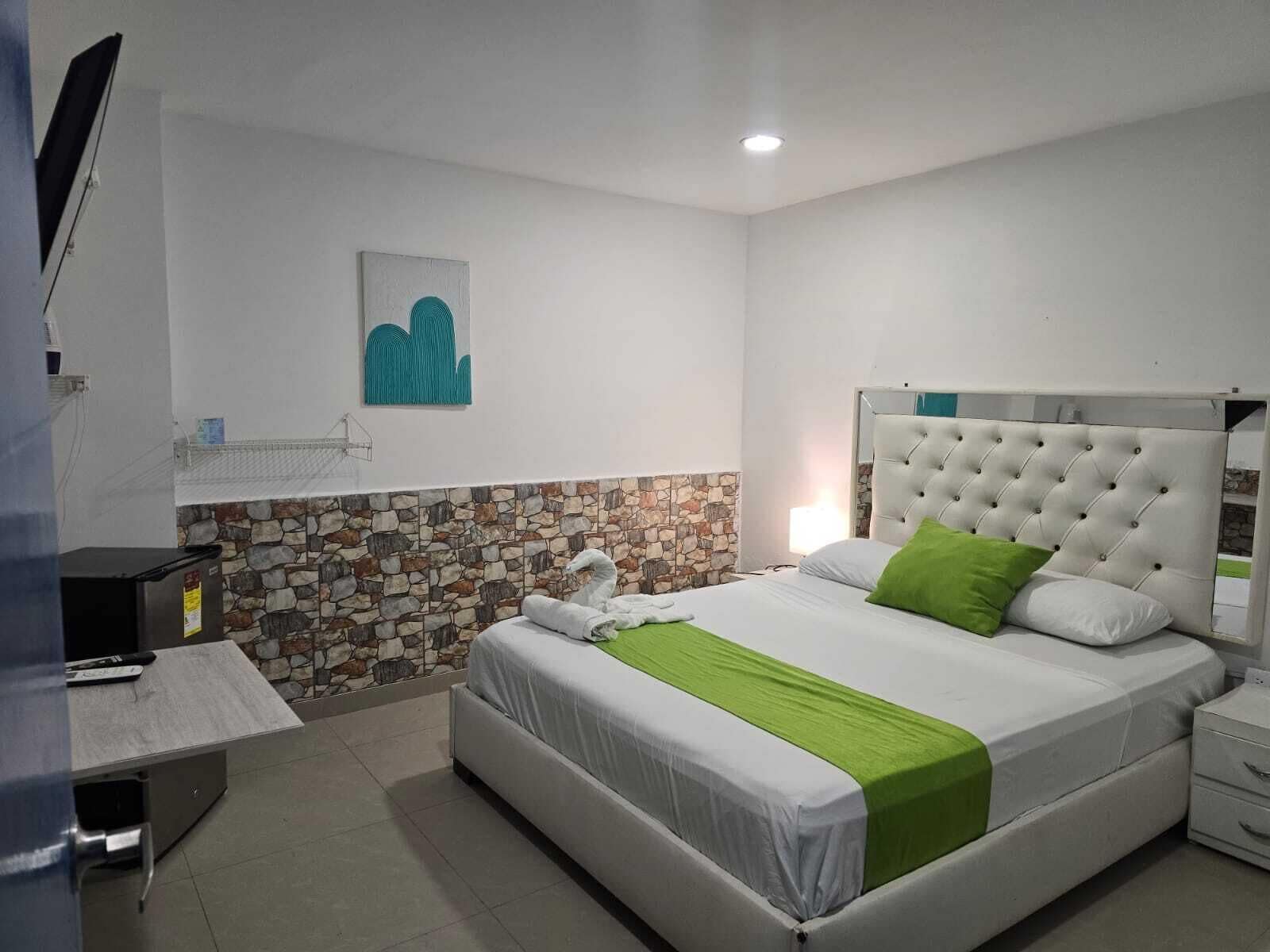 Ein Schlafzimmer mit Bett, Steinwand, weißem Kopfteil, grüner Decke, kleinem Tisch und Fernseher.