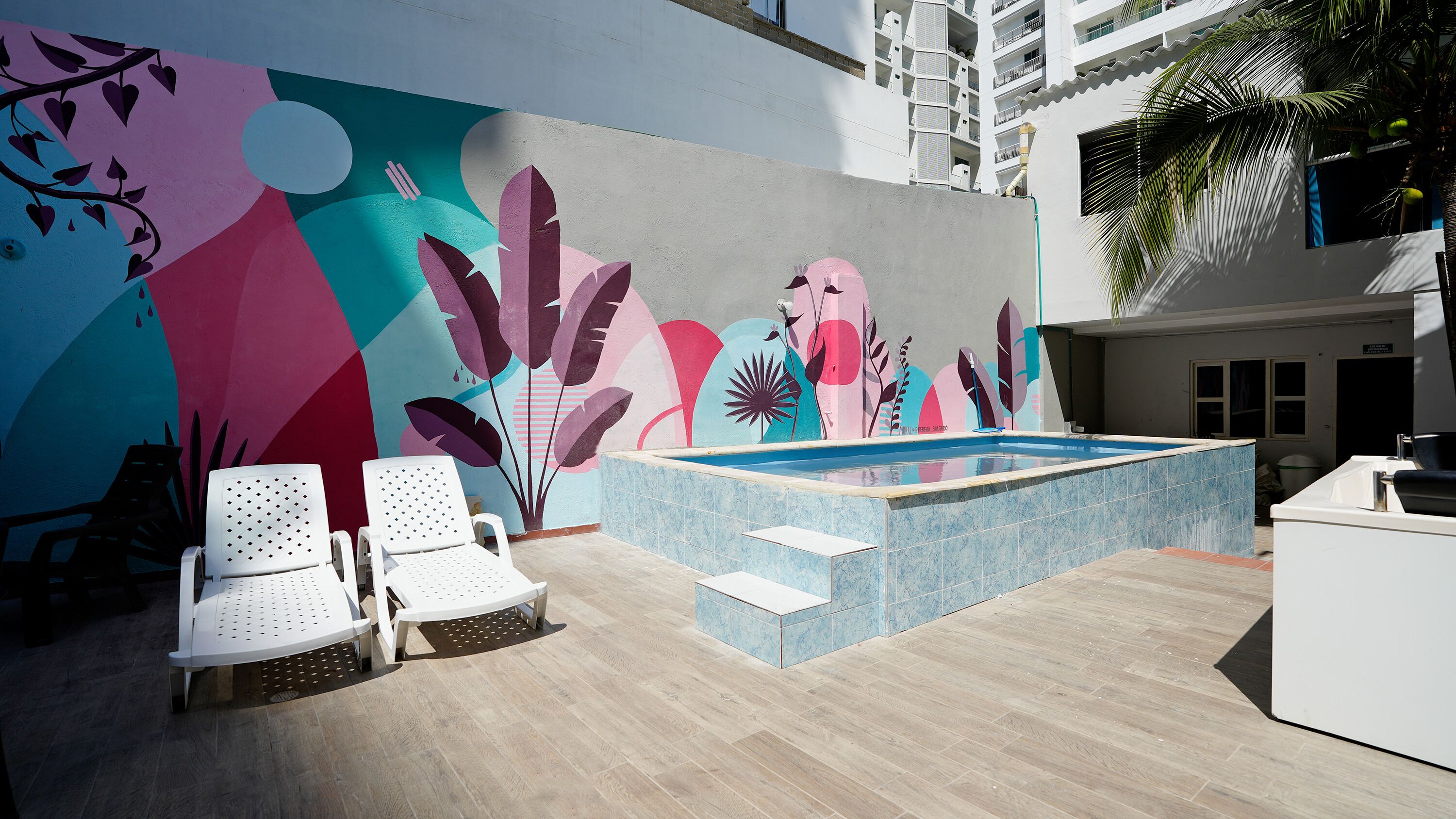 A modern outdoor pool area with a colorful mural, white lounge chairs, and a small hot tub.