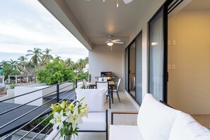 Un balcon avec un canapé blanc, une table à manger avec des chaises, un barbecue et une vue sur des palmiers.