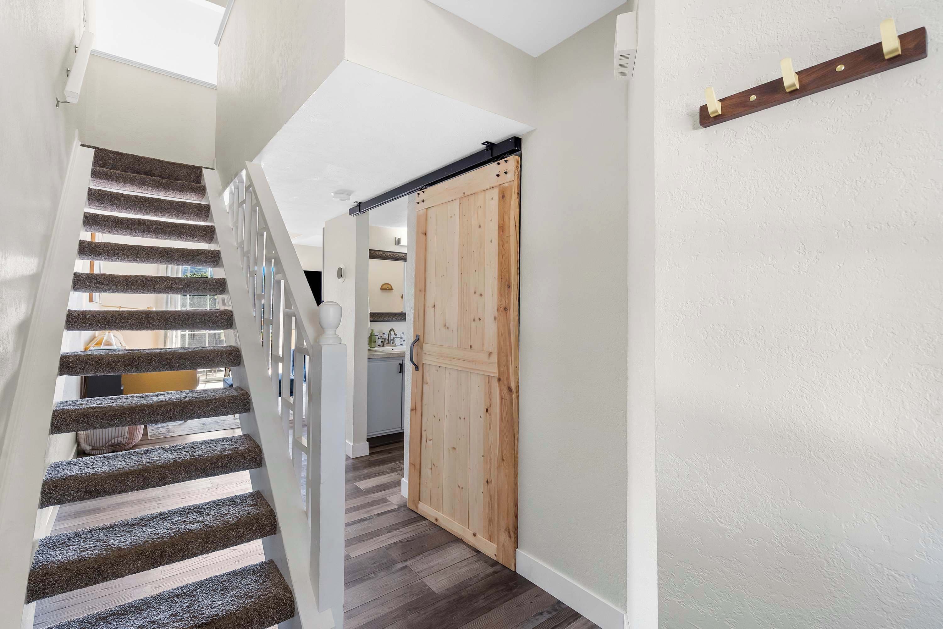 A staircase with a wooden finish and a white railing, leading to a room with a wooden door and a kitchen area visible in the background.