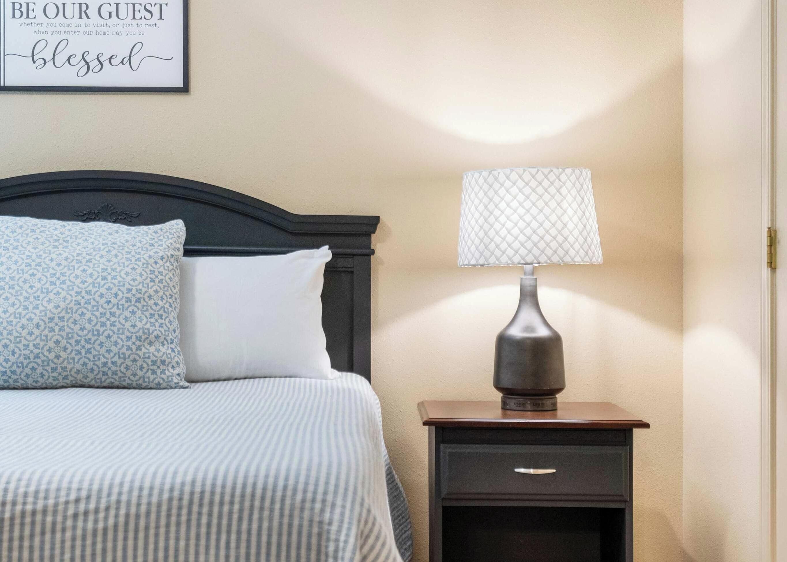 A neatly made bed with patterned pillows, a bedside table with a lamp, and a framed sign on the wall.