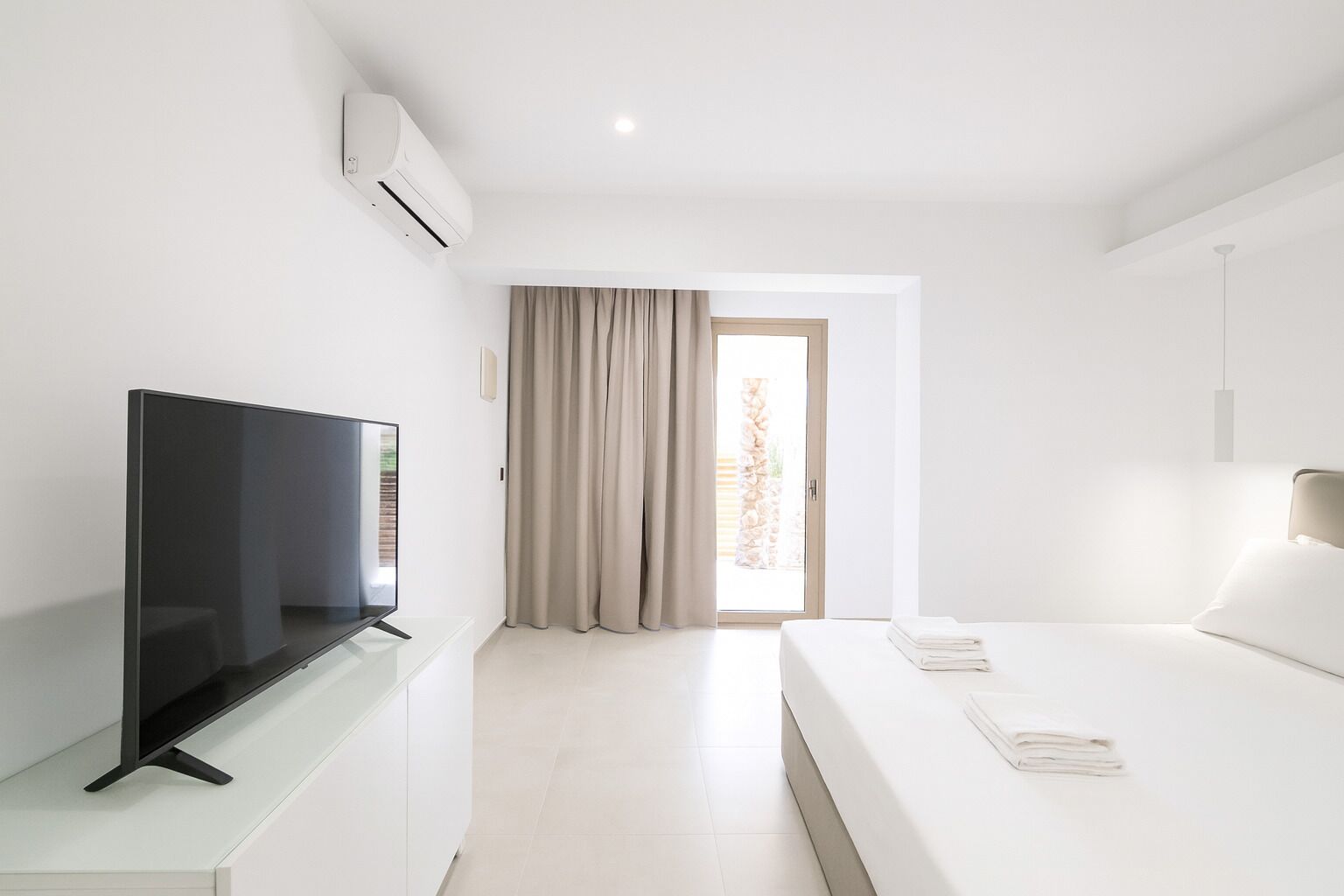 A modern hotel room with a flat-screen TV, a bed with white linens, a glass coffee table, and a large window with curtains.