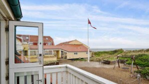 A beach house with a white balcony, a red flag, and a view of the sea and dunes.