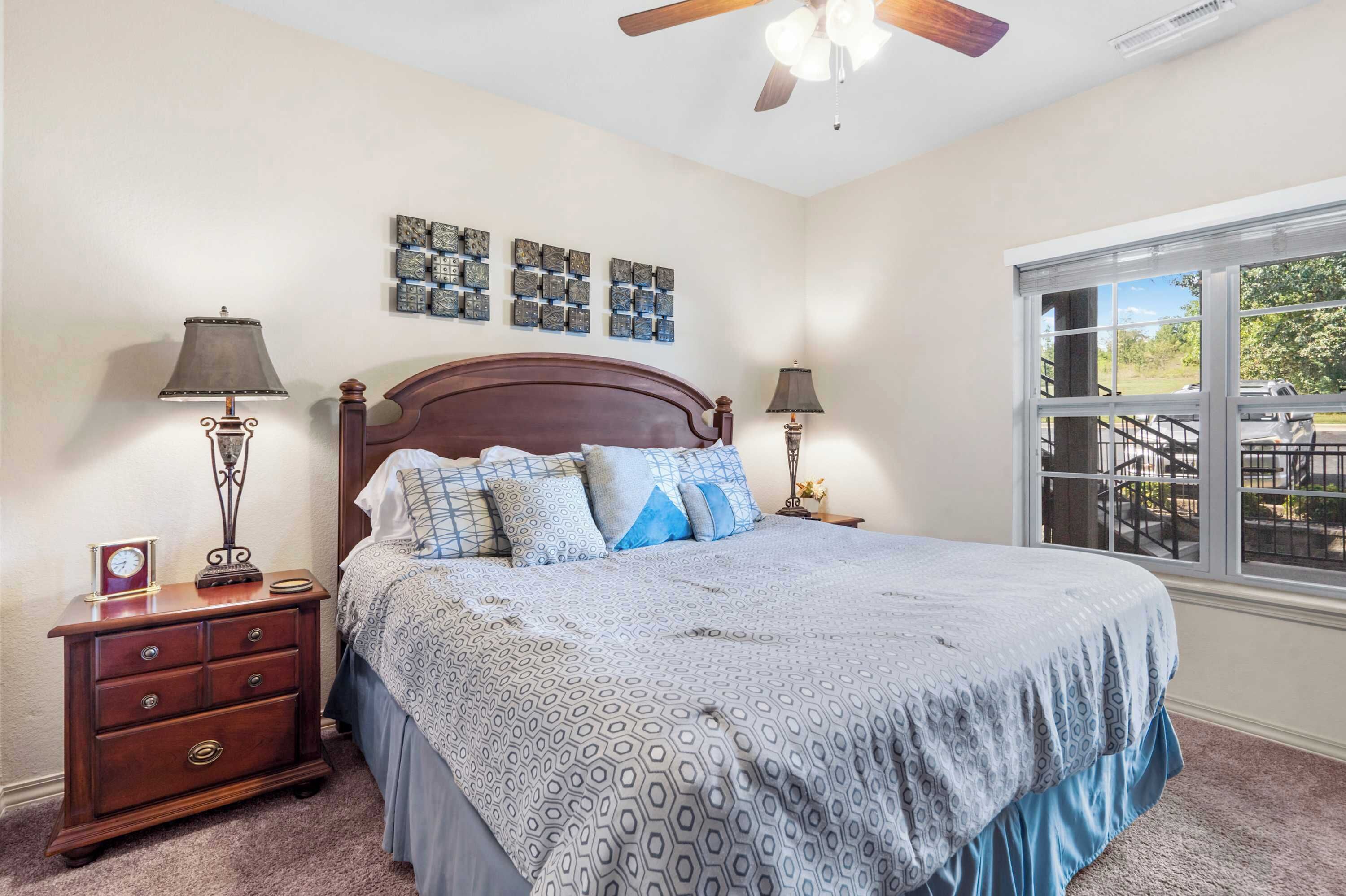 A bedroom with a large bed, two bedside tables, a ceiling fan, and a window with a view of the outdoors.