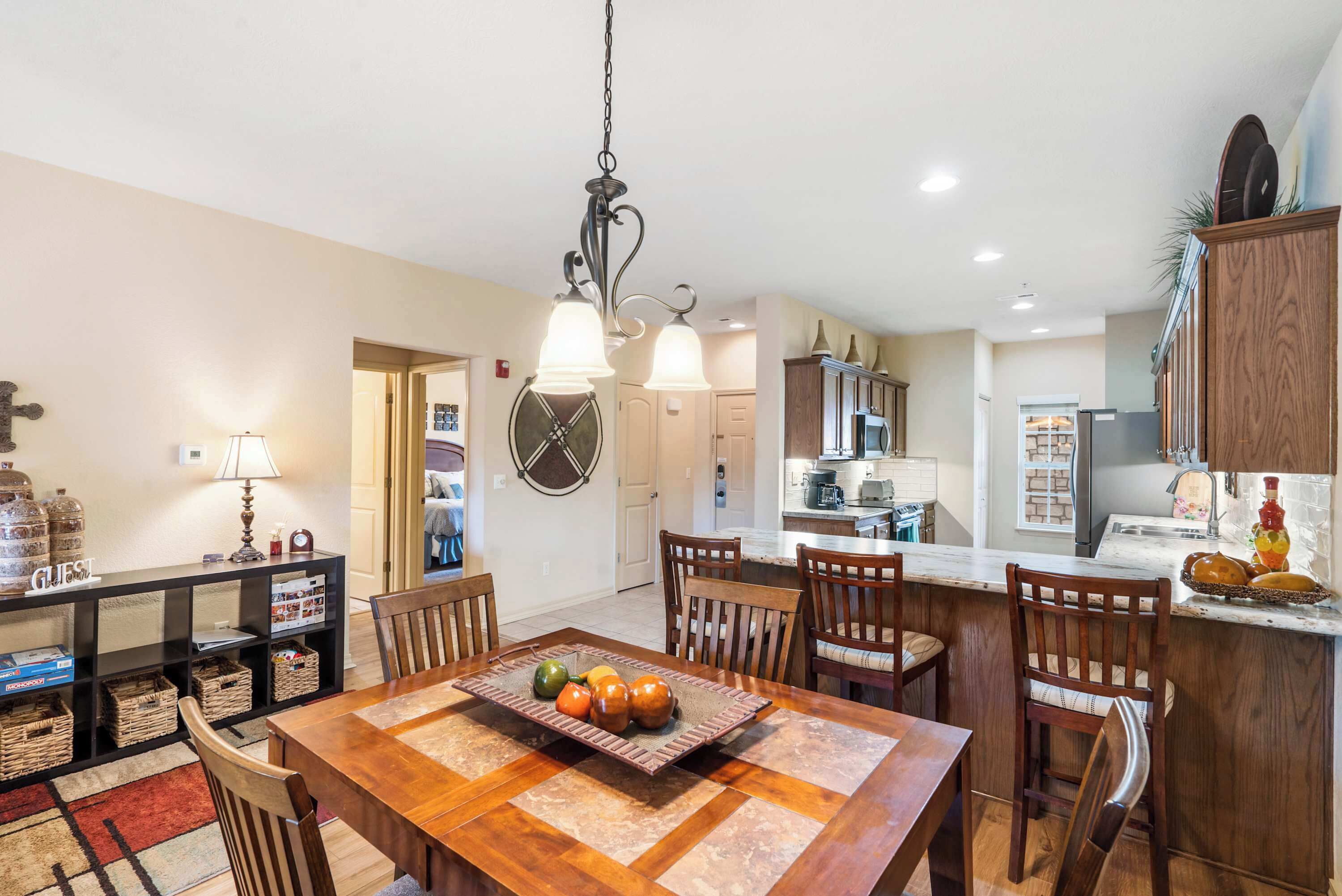 A dining area with a wooden table, chairs, and a fruit centerpiece. There is a kitchen in the background with cabinets and appliances.