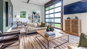 A modern living room with a sofa, coffee table, and a large window with blinds. - Historic 4-BR Loft near Ivy House | Saugatuck (Saugatuck)