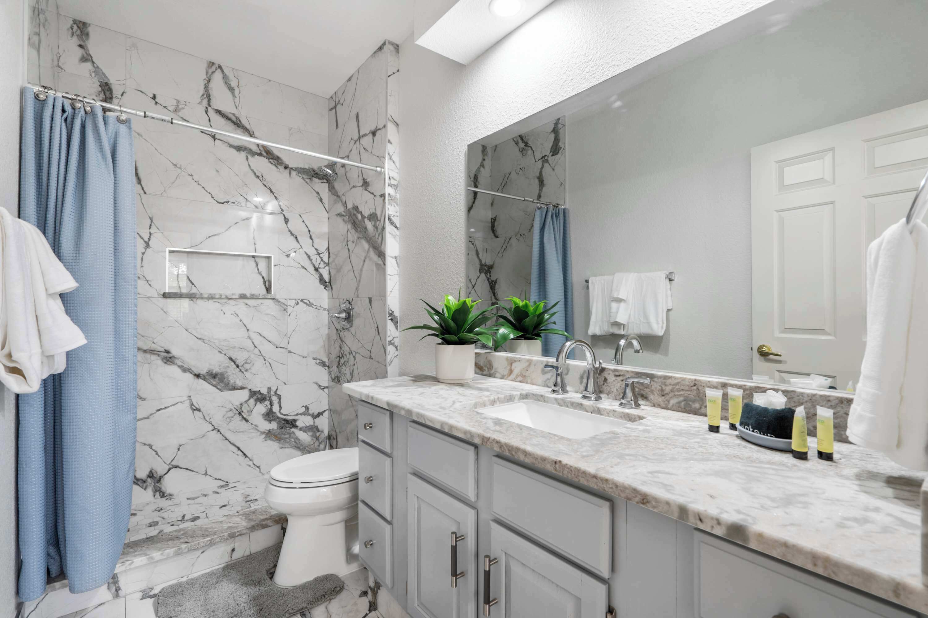 Une salle de bain aux murs recouverts de marbre, avec des toilettes blanches, un lavabo avec un miroir et des rideaux de douche bleus.