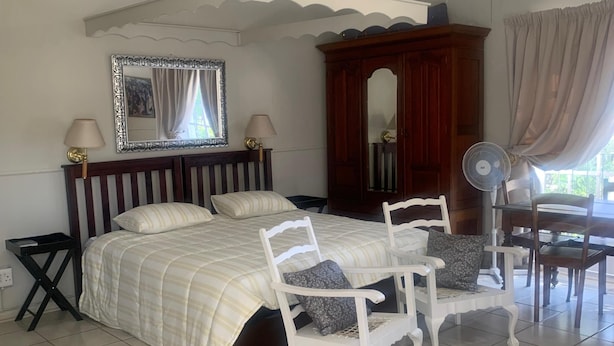 Ein Schlafzimmer mit einem Bett, zwei Nachttischen, einem Sessel, einem Schreibtisch, einem Ventilator, einem Deckenventilator, einem Spiegel und Vorhängen.