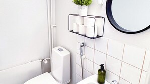 A modern bathroom with a toilet, sink, and bathtub. There is a toilet paper holder with a small plant, a round mirror, and a soap dispenser.