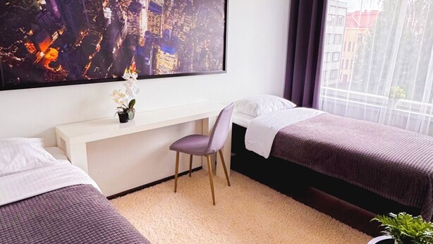 A hotel room with two beds, a desk, a large window, and a cityscape painting.