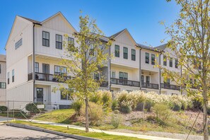 A multi-story residential building with balconies, a front porch, and a landscaped yard with various shrubs and trees.
