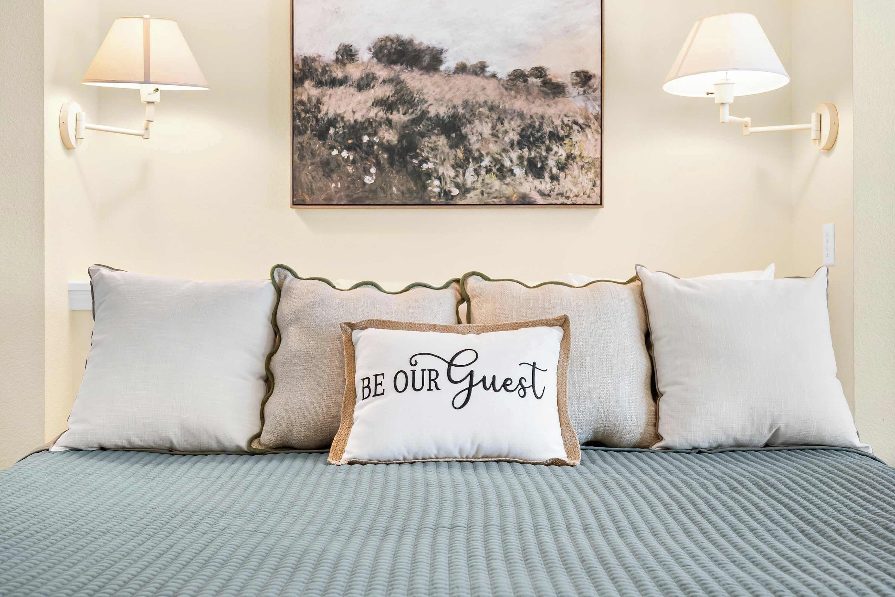 A neatly made bed with a textured grey bedspread, two wall-mounted lamps, and a painting of a landscape above the bed.