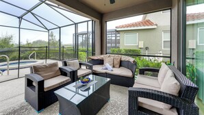 Un patio couvert aménagé avec du mobilier d’extérieur, une table en verre et une vue sur une piscine et la verdure.