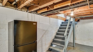 A basement with a stainless steel refrigerator, a concrete staircase, and exposed wooden beams.