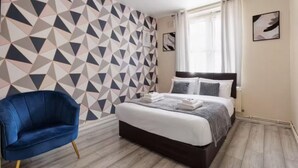 Una camera da letto con un letto, una poltrona blu, una parete con motivo geometrico e una finestra con tende.