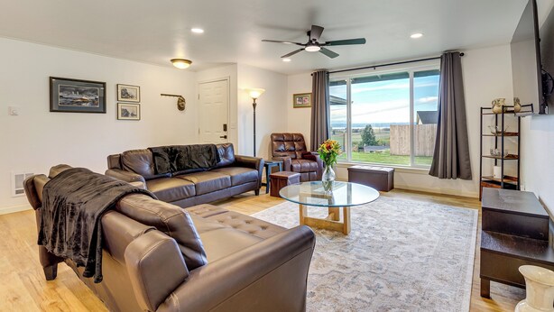 A living room with a leather sofa, a glass coffee table, and a view of the outdoors through large windows. - Explore Camano Island Beaches! Family Home w/ Yard (Stanwood)