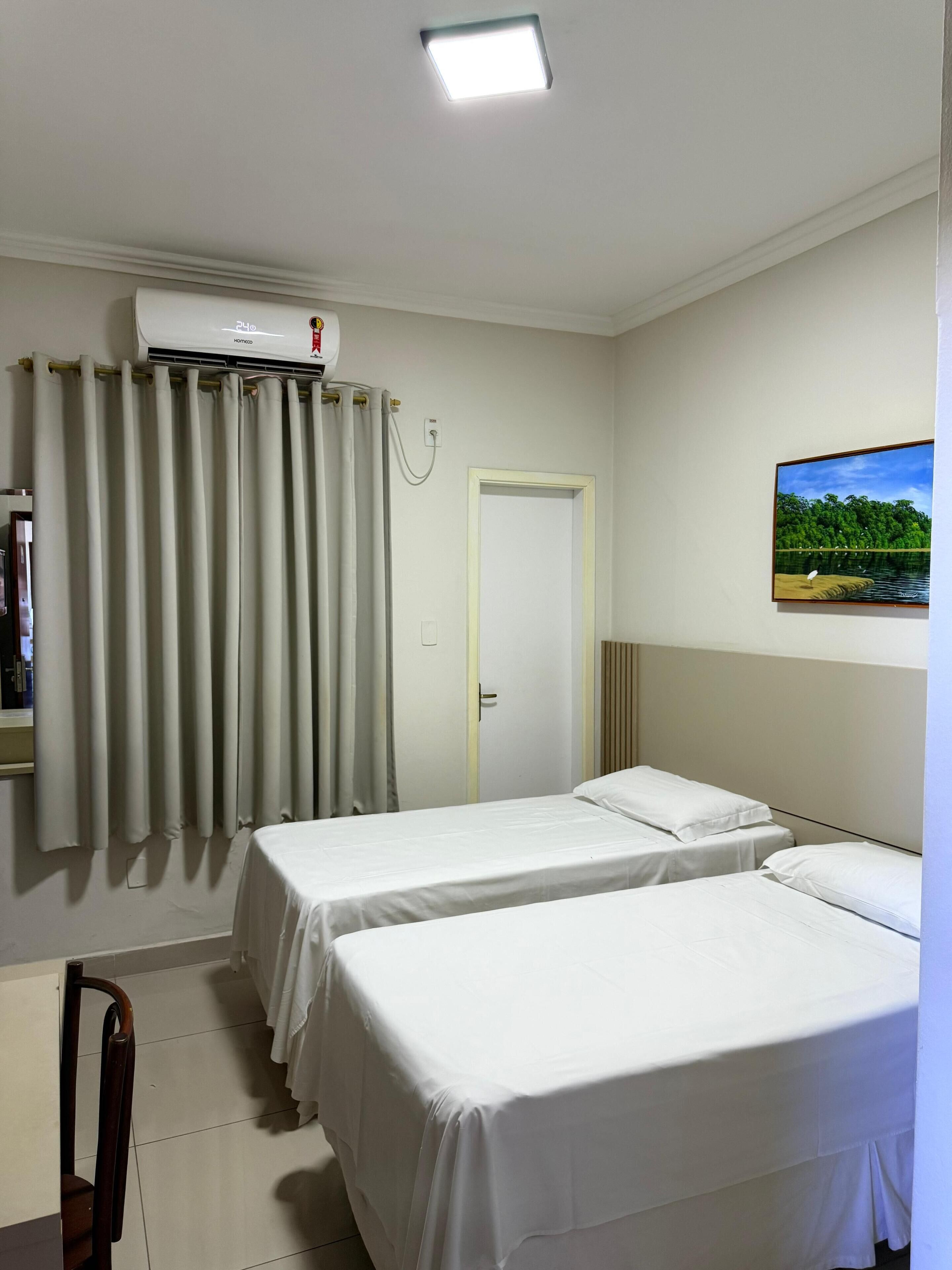 Ein Hotelzimmer mit zwei Betten, einem Vorhangfenster, einer Klimaanlage und einem Gemälde an der Wand.