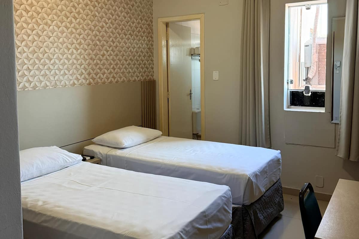 A hotel room with two beds, a window with curtains, an air conditioning unit, and a bathroom visible through an open door.
