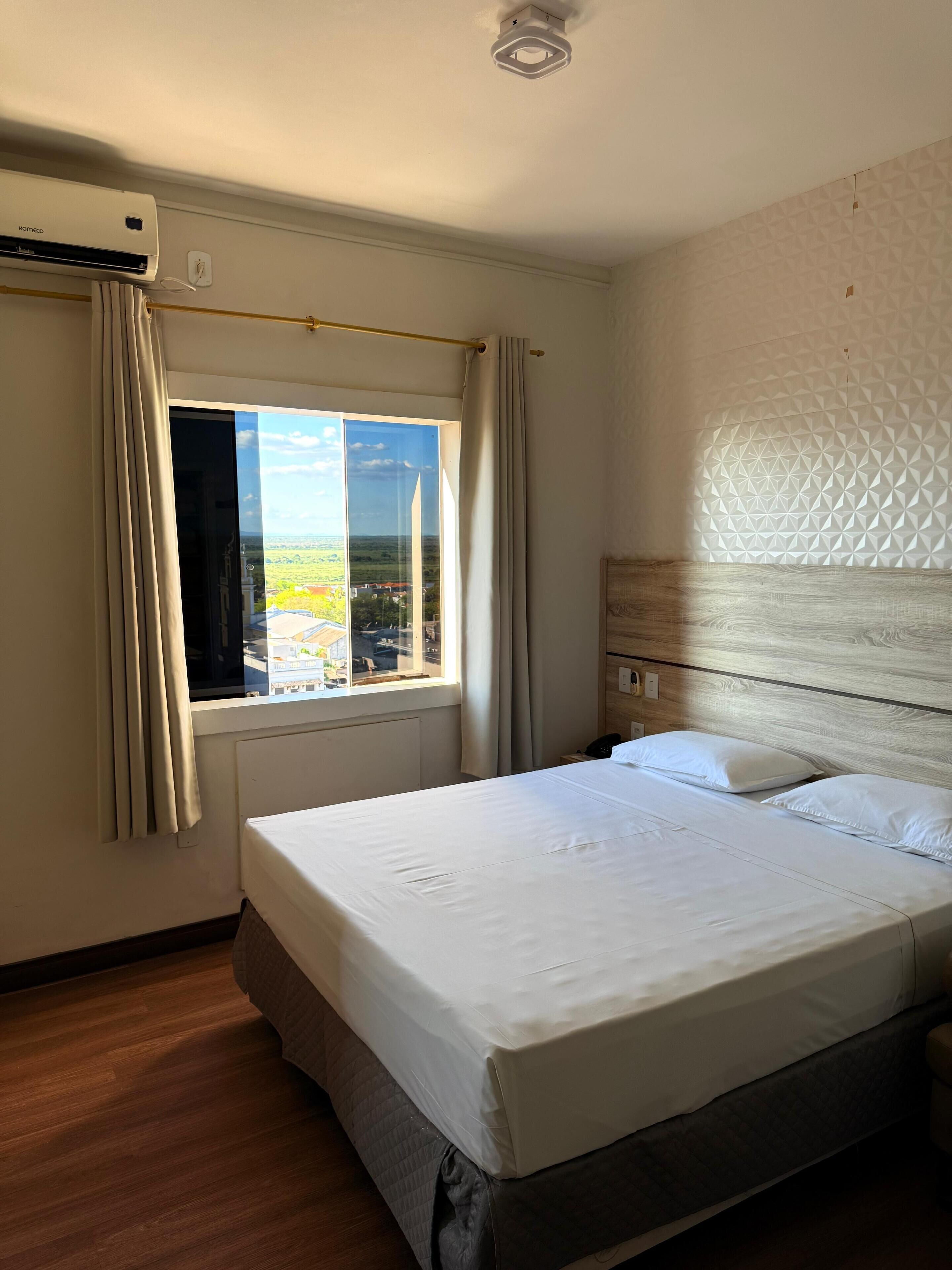 A hotel room with a large bed, a window with curtains, a ceiling light, and a view of the outside.