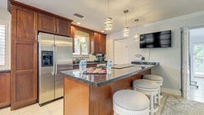 A modern kitchen with a stainless steel refrigerator, wooden cabinets, a granite countertop, and a built-in oven. There are three bar stools and a flat-screen TV mounted on the wall. - Walkable Island City Oasis w/ Private Heated Pool! (Wilton Manors)