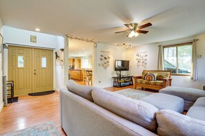 Ein geräumiges Wohnzimmer mit einem großen, grauen Ecksofa, einem Holztisch und einem Deckenventilator. Im Hintergrund ist eine Küche mit Holzschränken und einem Essbereich zu sehen.