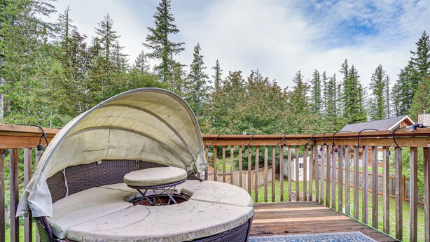 Un espace extérieur aménagé, abrité et doté d’un banc rembourré et d’une table, entouré d’une terrasse en bois et offrant une vue sur une zone boisée.